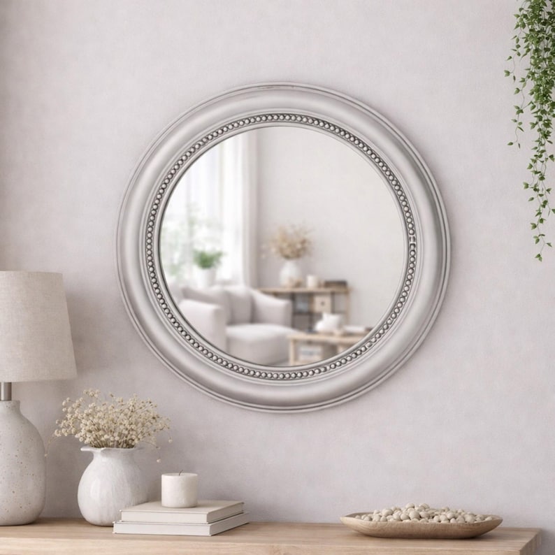 Pode incluir: Um espelho de parede redondo com uma moldura lisa cinza-clara, apresentando uma borda interna decorativa com contas. A superf&iacute;cie circular de vidro reflete um espa&ccedil;o interior luminoso e desfocado, capturando a luz e a profundidade do ambiente.