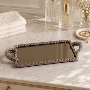 Peut inclure: Un plateau décoratif rectangulaire avec une surface miroir et des poignées en forme de corde. Le plateau est de couleur taupe clair et repose sur une surface en marbre. D'autres articles sur la surface comprennent des flacons de parfum, un petit vase de fleurs et des serviettes blanches pliées.