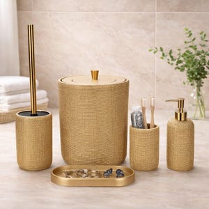 May include: A gold-coloured bathroom set with a textured finish. The set includes a toilet brush holder, a bin, a toothbrush holder, a soap dispenser, and a small tray. The items are arranged on a light-coloured surface.