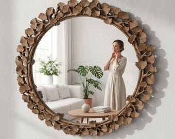 Op de afbeelding: Een ronde wandspiegel met een decoratieve lijst versierd met talloze gebeeldhouwde ginkgobladeren in een natuurlijke houttint. Het ronde glazen oppervlak weerspiegelt een kamerinterieur met een witte bank, een houten salontafel en een plant.