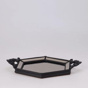 May include: Black hexagonal decorative tray with mirrored surfaces and ornate handles. The tray has a dark finish and is designed for displaying items. The handles have a detailed, floral-like design.
