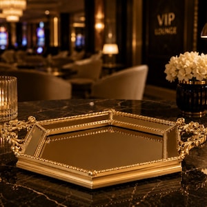 Può includere: Un vassoio a specchio esagonale dorato con manici decorati è appoggiato su un tavolo di marmo scuro. Una candela in un portacandele di vetro e un vaso di fiori bianchi sono anche sul tavolo. Lo sfondo è una VIP lounge scarsamente illuminata.