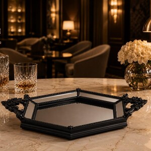 May include: A black decorative tray with a mirrored surface and ornate handles sits on a marble table. A crystal decanter and glass with amber liquid are to the left, and a vase of white flowers is to the right.