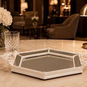May include: A white, mirrored tray with floral handles sits on a marble table. The tray has a geometric shape with a mirrored surface and a white frame. A glass and a vase of white flowers are also on the table.
