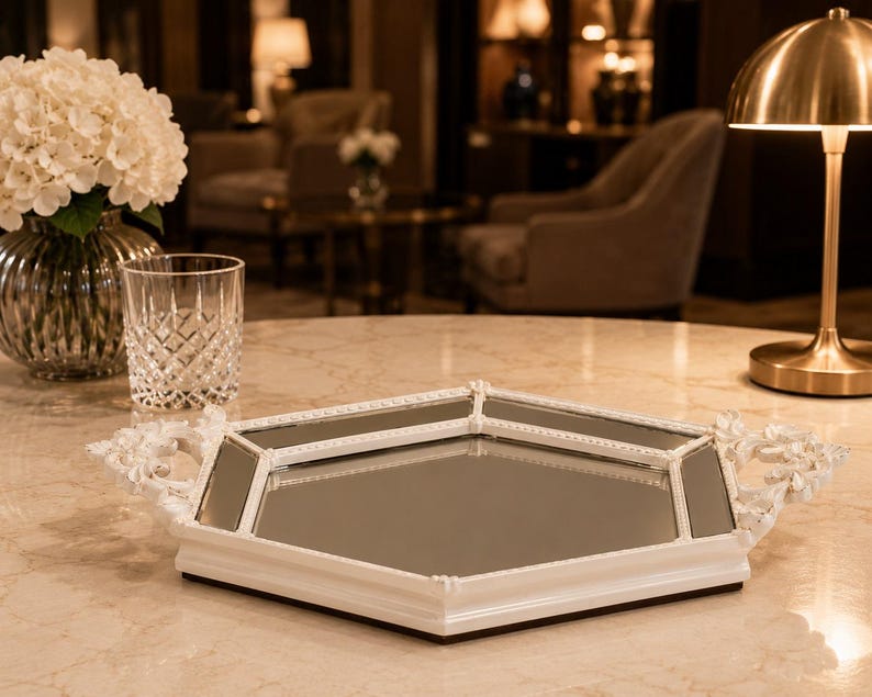 May include: A white, mirrored tray with floral handles sits on a marble table. The tray has a geometric shape with a mirrored surface and a white frame. A glass and a vase of white flowers are also on the table.