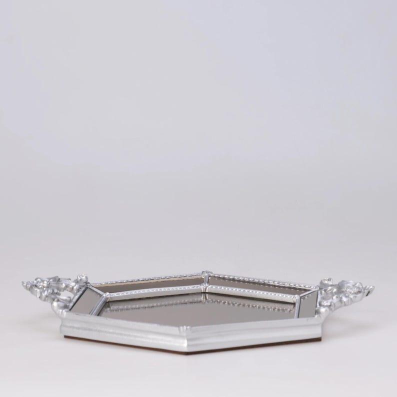 May include: A silver-colored, geometric tray with a mirrored surface. The tray has decorative handles on each side and a brown base. The tray is designed with a modern aesthetic.