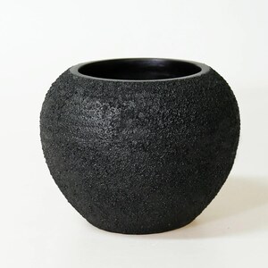 Peut inclure: Un vase décoratif noir de forme arrondie avec une surface texturée et rugueuse. Le vase présente une large ouverture et une forme sphérique, idéal pour exposer des fleurs ou comme pièce d'art. La couleur sombre et la texture unique ajoutent une touche moderne.