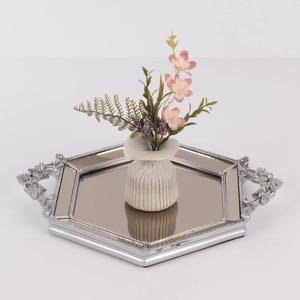 May include: A decorative silver-toned tray with a mirrored surface and ornate handles. A small, textured vase holds a floral arrangement with pink flowers and green foliage. The tray is hexagonal in shape and sits on a white surface.