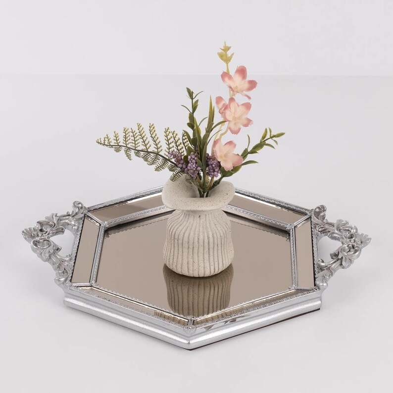 May include: A decorative silver-toned tray with a mirrored surface and ornate handles. A small, textured vase holds a floral arrangement with pink flowers and green foliage. The tray is hexagonal in shape and sits on a white surface.