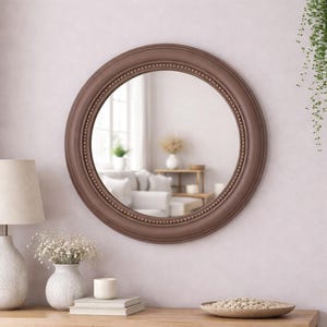 Pode incluir: Um espelho de parede redondo com uma moldura castanha mate com um detalhe de rebordo interior com contas. A superf&iacute;cie circular de vidro reflete uma sala interior luminosa com um sof&aacute;, uma mesa de madeira e um vaso branco.
