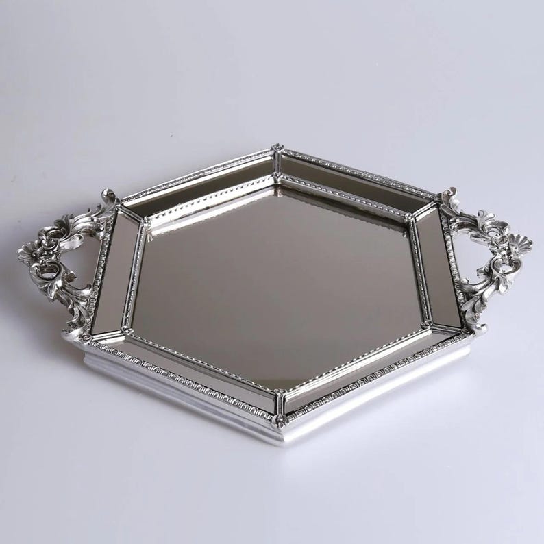 May include: A silver-colored, hexagonal serving tray with ornate handles. The tray has a mirrored surface and a decorative border. The handles feature a floral design. The tray is approximately 16 inches long and 12 inches wide.