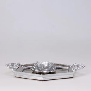 May include: A silver-colored decorative tray with a mirrored surface and a silver flower-shaped centerpiece. The tray has a geometric shape with ornate handles on each side. The tray is likely used for decorative purposes.