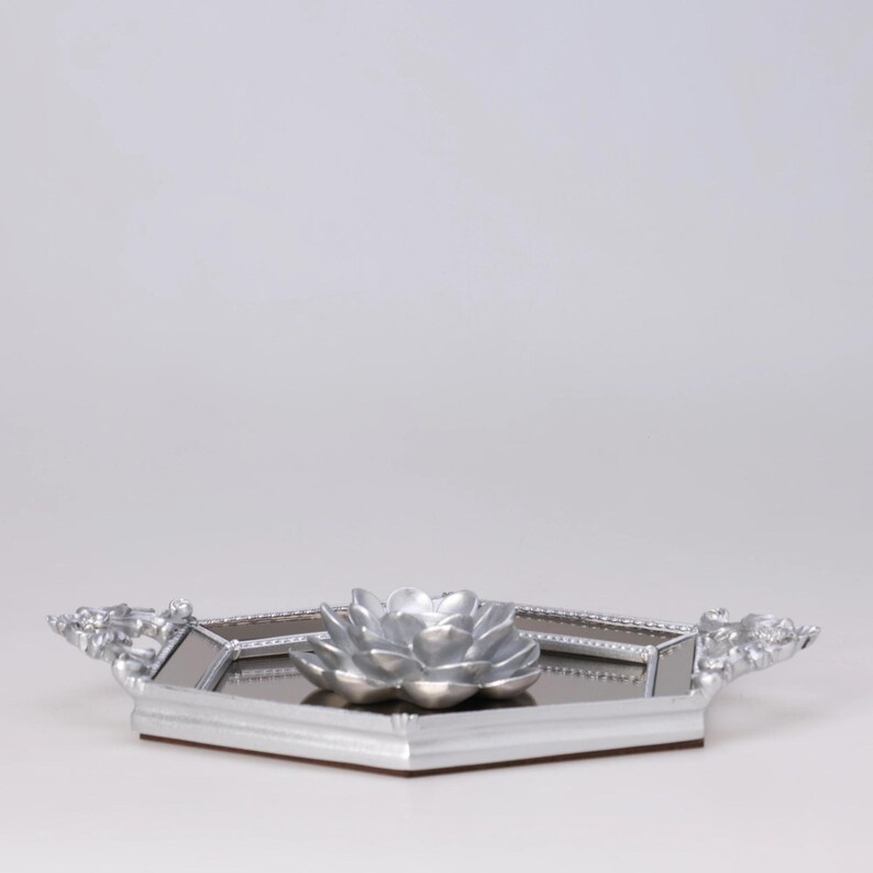 May include: A silver-colored decorative tray with a mirrored surface and a silver flower-shaped centerpiece. The tray has a geometric shape with ornate handles on each side. The tray is likely used for decorative purposes.