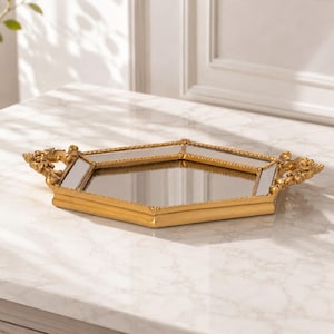 May include: A hexagonal mirrored tray with a gold-toned frame. The tray features a beaded edge detail and ornate, decorative handles on two sides. The central surface is a flat, reflective mirror, and the frame has a smooth, metallic finish.