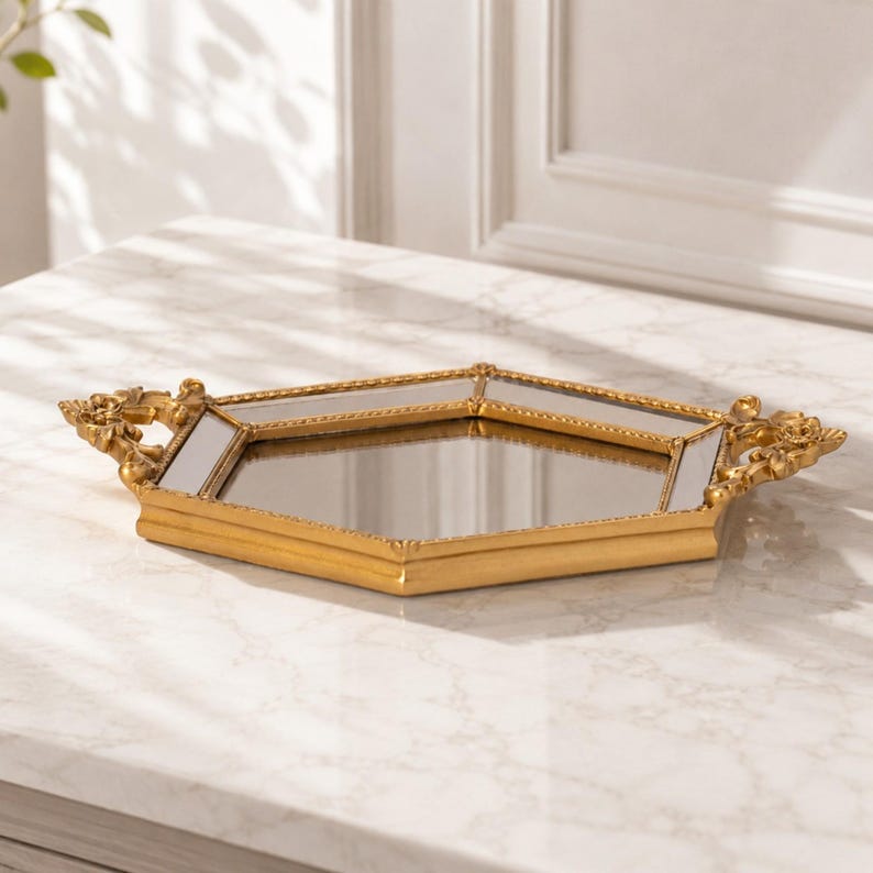 May include: A hexagonal mirrored tray with a gold-toned frame. The tray features a beaded edge detail and ornate, decorative handles on two sides. The central surface is a flat, reflective mirror, and the frame has a smooth, metallic finish.