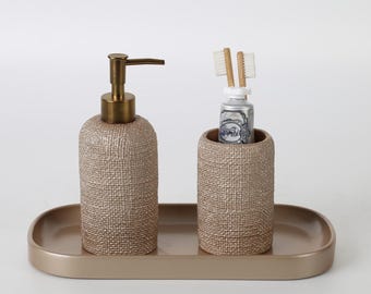 May include: A bathroom set featuring a beige textured soap dispenser with a bronze pump, a matching cylindrical toothbrush holder, and an oval tray. The toothbrush holder contains two wooden toothbrushes and a silver tube.