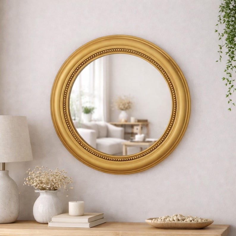 Pode incluir: Um espelho de parede redondo com uma moldura dourada larga e lisa, apresentando uma borda interna decorativa de contas. A superf&iacute;cie circular de vidro reflete um espa&ccedil;o interior luminoso, capturando a luz e a profundidade da sala.