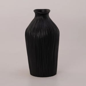 Könnte beinhalten: Eine hohe, schwarze Vase mit einer strukturierten, vertikal geriffelten Oberfläche. Die Vase hat eine leicht ausgestellte Öffnung und einen abgerundeten Boden. Ein vielseitiges Dekorationsobjekt für jedes Zuhause.