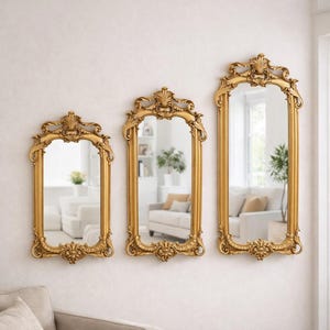 May include: Three ornate, gold-framed mirrors of varying sizes are mounted on a white wall. Each mirror has a decorative, scrolled design at the top and bottom. The mirrors reflect a room with white furniture and plants.