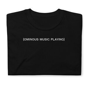 May include: Black t-shirt with the text "{OMINOUS MUSIC PLAYING}" printed in white. The shirt is made of a soft material and has a classic crew neck design. The text is centered on the front of the shirt.