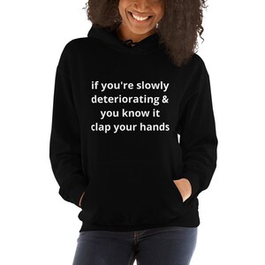 May include: Black hooded sweatshirt with the text "if you're slowly deteriorating & you know it clap your hands" in white. The sweatshirt has a front pocket and a drawstring hood. The model is wearing the sweatshirt.