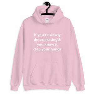 May include: A light pink hoodie with a front pocket and a drawstring hood. The hoodie has the text "if you're slowly deteriorating & you know it clap your hands" printed in white.
