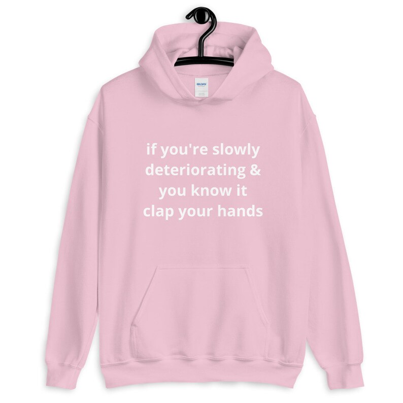 May include: A light pink hoodie with a front pocket and a drawstring hood. The hoodie has the text "if you're slowly deteriorating & you know it clap your hands" printed in white.
