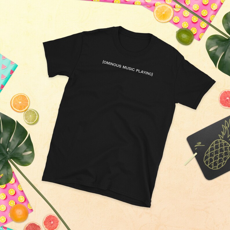 May include: Black t-shirt with the text "[OMINOUS MUSIC PLAYING]" in white. The shirt is short-sleeved and has a crew neck. The background includes tropical fruit and foliage.