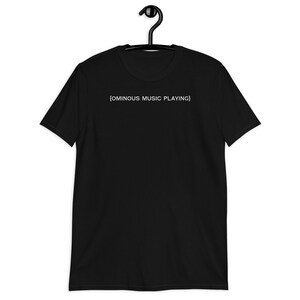 May include: Black short-sleeve t-shirt hanging on a black hanger. The shirt has the text "{OMINOUS MUSIC PLAYING}" printed in white across the chest. The t-shirt is made of a soft material.
