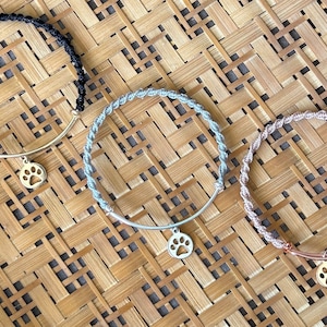 May include: Three bracelets with a paw print charm. The bracelets are made of black, gray, and rose gold cord. The charms are made of gold metal.