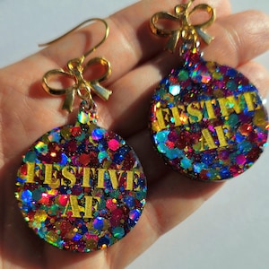 May include: A pair of festive earrings with a gold bow and hook. The round, glitter-filled ornaments feature the text "FESTIVE AF" in gold. The earrings are a mix of red, blue, green, and pink glitter.