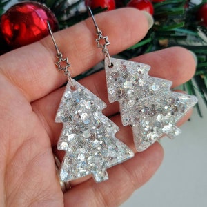 Könnte beinhalten: Silberne Glitzer-Weihnachtsbaum-Ohrringe mit Stern-Akzenten. Die Ohrringe sind wie Weihnachtsbäume geformt und mit silbernem Glitzer gefüllt. Sie hängen an silbernen Haken mit einem kleinen Stern.