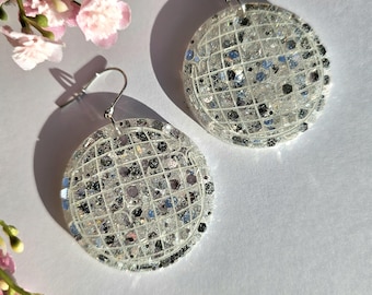 May include: A pair of silver-toned dangle earrings. Each earring is a clear, circular disc filled with silver and black glitter and a grid pattern. The earrings have a hook closure.