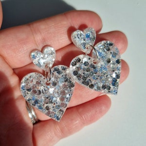 May include: A pair of dangle earrings featuring two heart-shaped charms each. The smaller heart sits above a larger heart, both crafted from clear material embedded with chunky silver and holographic glitter that reflects light with a metallic finish.