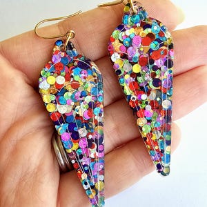 Könnte beinhalten: Ein Paar farbenfrohe, glitzergefüllte Ohrringe. Die Ohrringe haben eine tropfenförmige Form mit einem goldfarbenen Haken. Die Ohrringe sind mit einer Vielzahl von bunten Pailletten gefüllt, darunter Rot, Gelb, Blau und Lila.