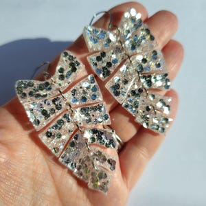 May include: A pair of silver glitter leaf earrings. The earrings are made of clear resin and have a silver glitter finish. The earrings are shaped like leaves and have a dangling design.