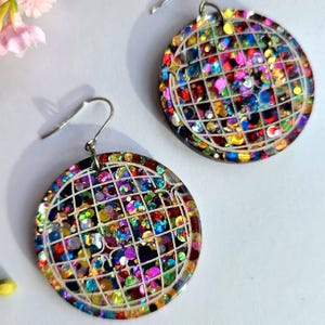 May include: A pair of round, colourful earrings with a disco ball design. The earrings feature a grid pattern over a background of multicoloured sequins. The earrings have silver-coloured ear wires.