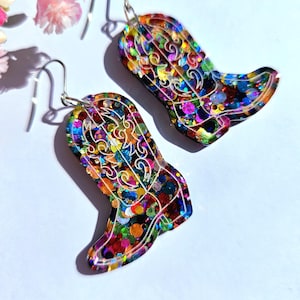 May include: A pair of colourful, glittery cowboy boot earrings. The earrings are shaped like boots with intricate white filigree designs. The boots are filled with a mix of vibrant colours, including red, blue, green, and yellow, and are suspended from silver hooks.