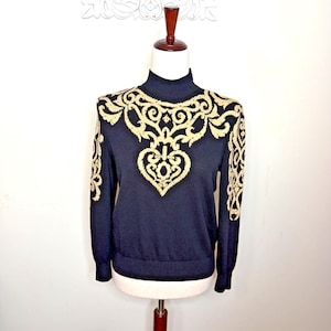 Op de afbeelding: Een marineblauwe coltrui met goudkleurige geborduurde details. Het sierlijke ontwerp heeft een hartvormig motief in het midden en loopt over de schouders en mouwen. De trui wordt op een mannequin gepresenteerd.