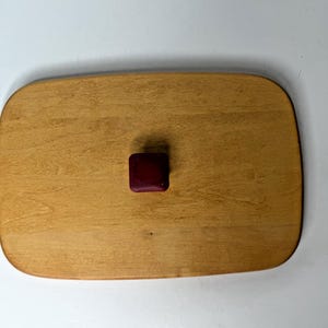 May include: A rectangular wooden lid with rounded corners and a smooth, light brown grain finish. Centered on the top surface is a small, square, dark maroon knob with a matte texture, providing a contrast to the natural wood grain of the lid.