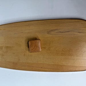 May include: A light brown wooden serving board with a smooth, elongated oval shape and rounded edges. The board features a small, square wooden handle centered on the top surface. The wood grain is visible across the entire surface of the board.