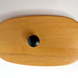 May include: An oval-shaped light brown wooden lid featuring a smooth, polished surface and a centered, small, round black knob handle. The wood grain is visible across the surface, and the edges of the lid are rounded for a finished appearance.