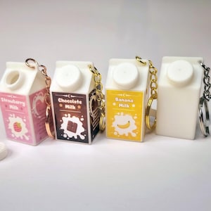 Könnte beinhalten: Vier Miniatur-Schlüsselanhänger in Form von Milchkartons, in den Geschmacksrichtungen Erdbeere, Schokolade, Banane und Naturweiß. Jeder Karton hat einen weißen Deckel und eine Metallkette mit einem Schlüsselring. Der Erdbeer-Karton hat einen rosa Hintergrund mit einer Erdbeer-Grafik.