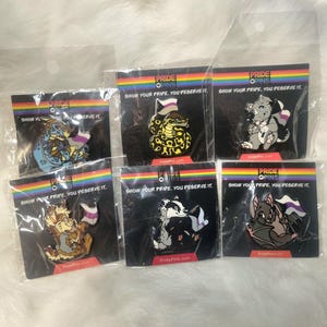 May include: Six enamel pins featuring different animals and a pride flag. The pins are all black and white with a rainbow pride flag and a purple, white and black asexual pride flag. The animals include a snake, a kangaroo, a dragon, a badger, a bat, and a cat. The text on the pins reads "Show your pride, you deserve it."