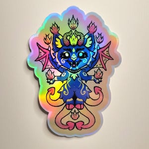 Peut inclure: Autocollant holographique représentant une créature bleue de type chat avec des flammes, des cornes et des ailes. L'autocollant a une bordure irisée colorée et un design fantaisiste. La créature a une expression amicale et est décorée de volutes et de motifs.