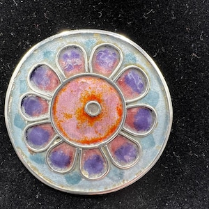 May include: A circular pendant with a floral pattern, featuring a silver frame. The centre is pink and orange, with purple and blue petal shapes around it. The pendant is set against a black backdrop.
