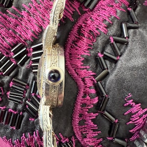 May include: Close-up of a silver-toned watch with an ornate band and a round face. The watch face features a dark blue stone. The watch is set against a background of black fabric with pink embroidery and black beads.