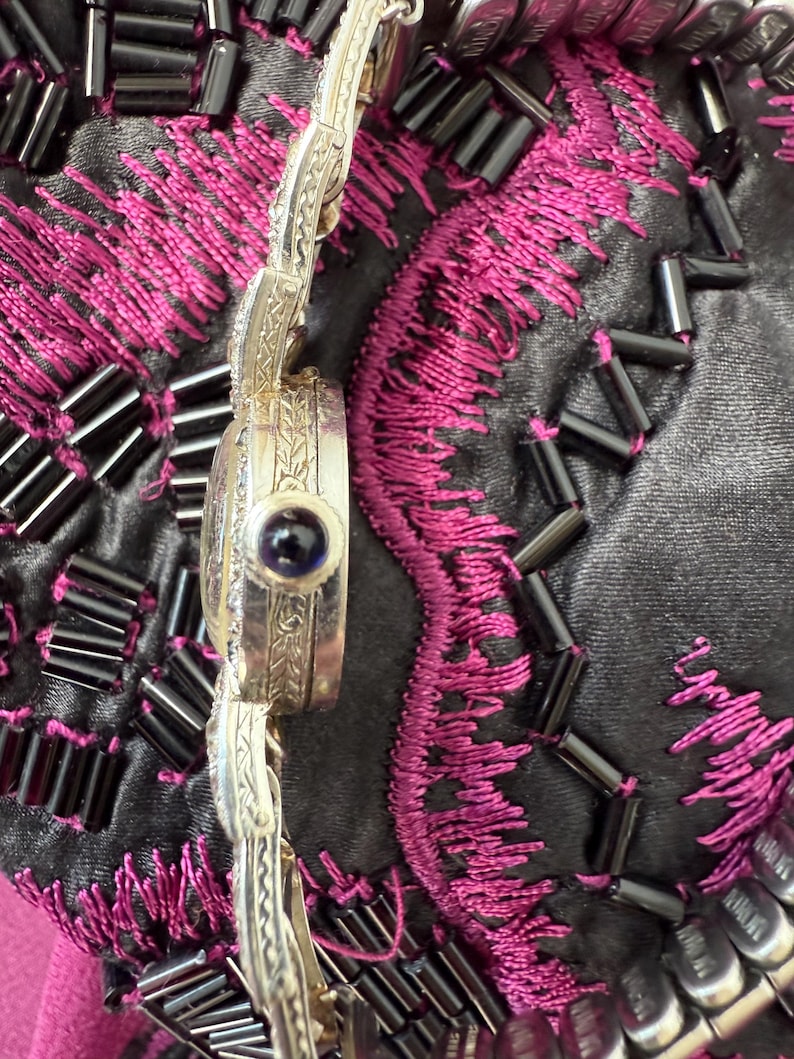 May include: Close-up of a silver-toned watch with an ornate band and a round face. The watch face features a dark blue stone. The watch is set against a background of black fabric with pink embroidery and black beads.