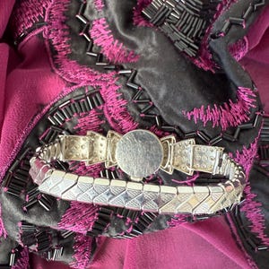 May include: A silver-toned bracelet with a geometric design, featuring a round central element. The bracelet is set against a backdrop of a dark gray fabric with magenta embroidery and a magenta fabric.