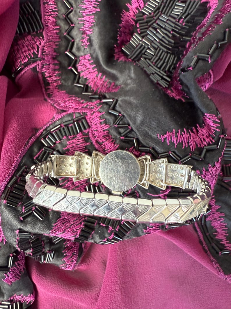May include: A silver-toned bracelet with a geometric design, featuring a round central element. The bracelet is set against a backdrop of a dark gray fabric with magenta embroidery and a magenta fabric.